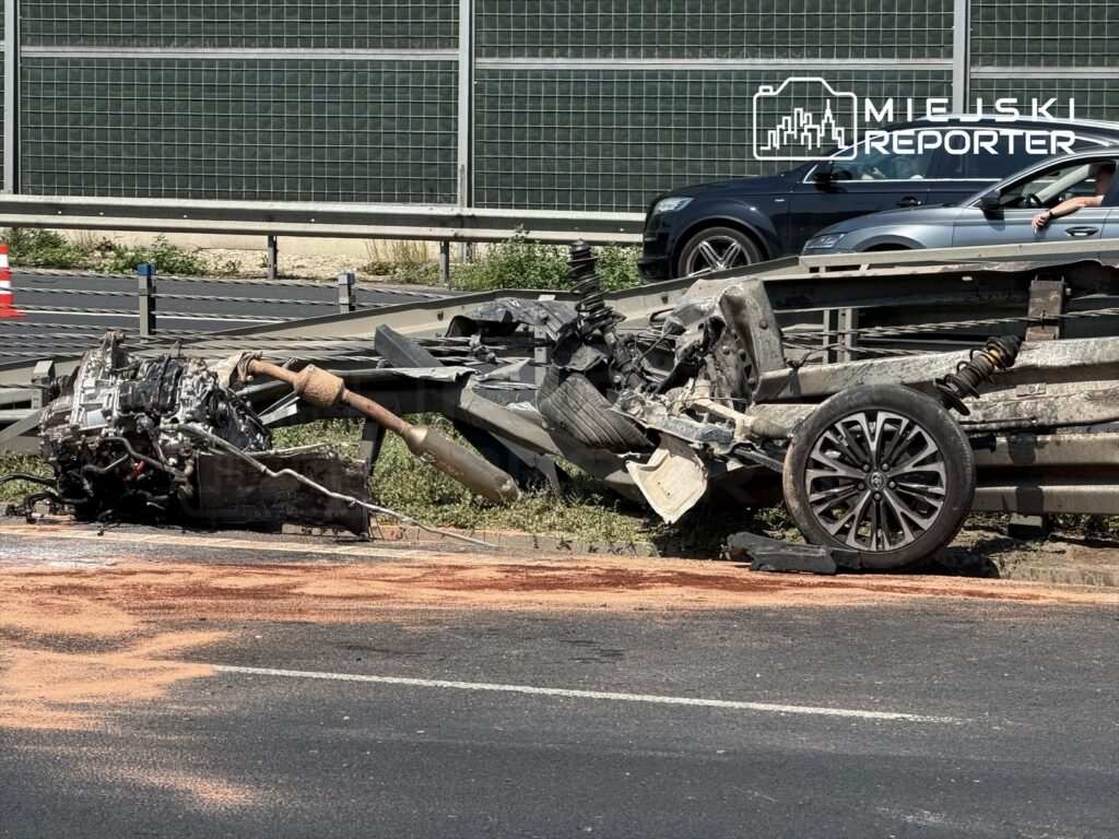 Uszkodzony silnik i elementy zawieszenia leżą na jezdni obok przewróconego pojazdu, przy barierkach drogi.