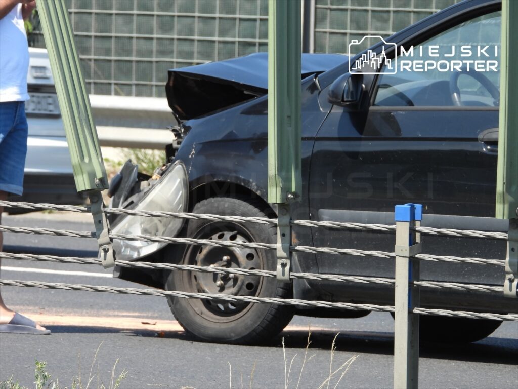 Czarny samochód osobowy z uszkodzoną przednią częścią stoi na drodze, obok mężczyzna w niebieskich spodniach rozmawia przez telefon.
