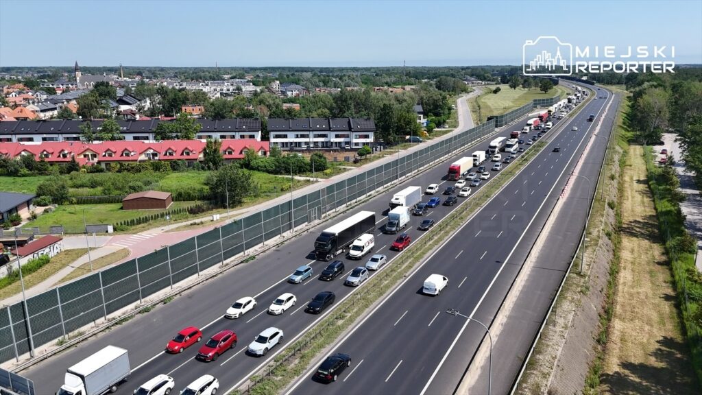 Zgromadzenie pojazdów na ruchliwej drodze ekspresowej, otoczone nowoczesnymi budynkami mieszkalnymi i zielenią.