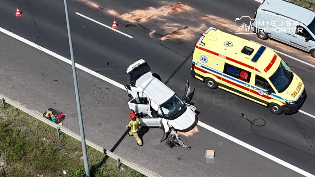 Na drodze leży uszkodzone auto osobowe z otwartymi drzwiami, obok stoi strażak w hełmie, a w tle widoczna karetka pogotowia.