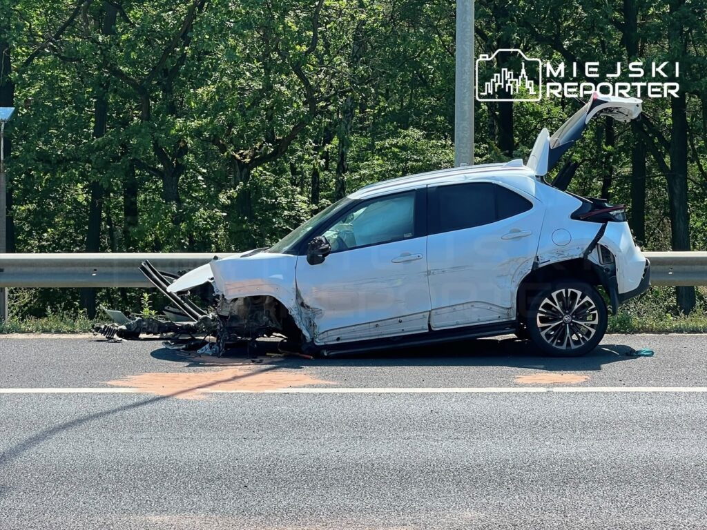 Uszkodzony samochód osobowy stoi na poboczu drogi, z otwartym bagażnikiem i usuniętymi elementami podwozia.