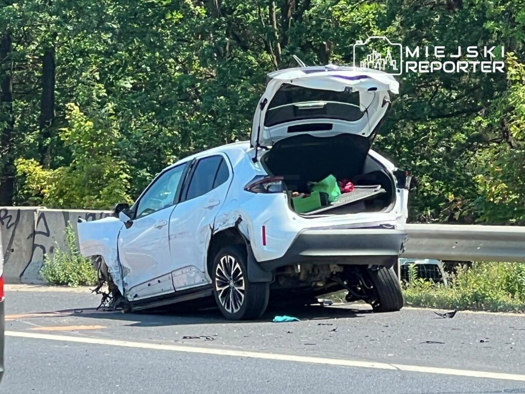 Uszkodzona biała Toyota z otwartym bagażnikiem stoi na poboczu drogi, obok widoczne fragmenty pojazdu.