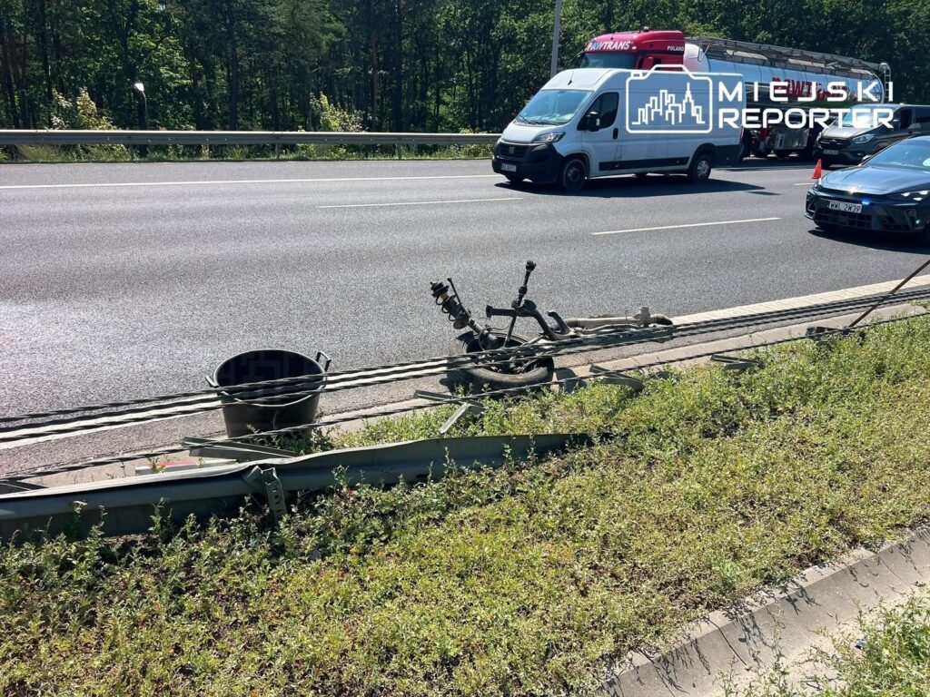 Na poboczu drogi leży silnik oraz koła, obok znajduje się czarna wiadro i fragmenty metalowych elementów. W tle widać ruch samochodów.