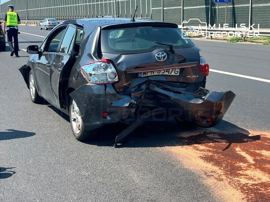 Czarny samochód osobowy z uszkodzoną tylną częścią stoi na drodze, obok widać policjanta prowadzącego czynności.