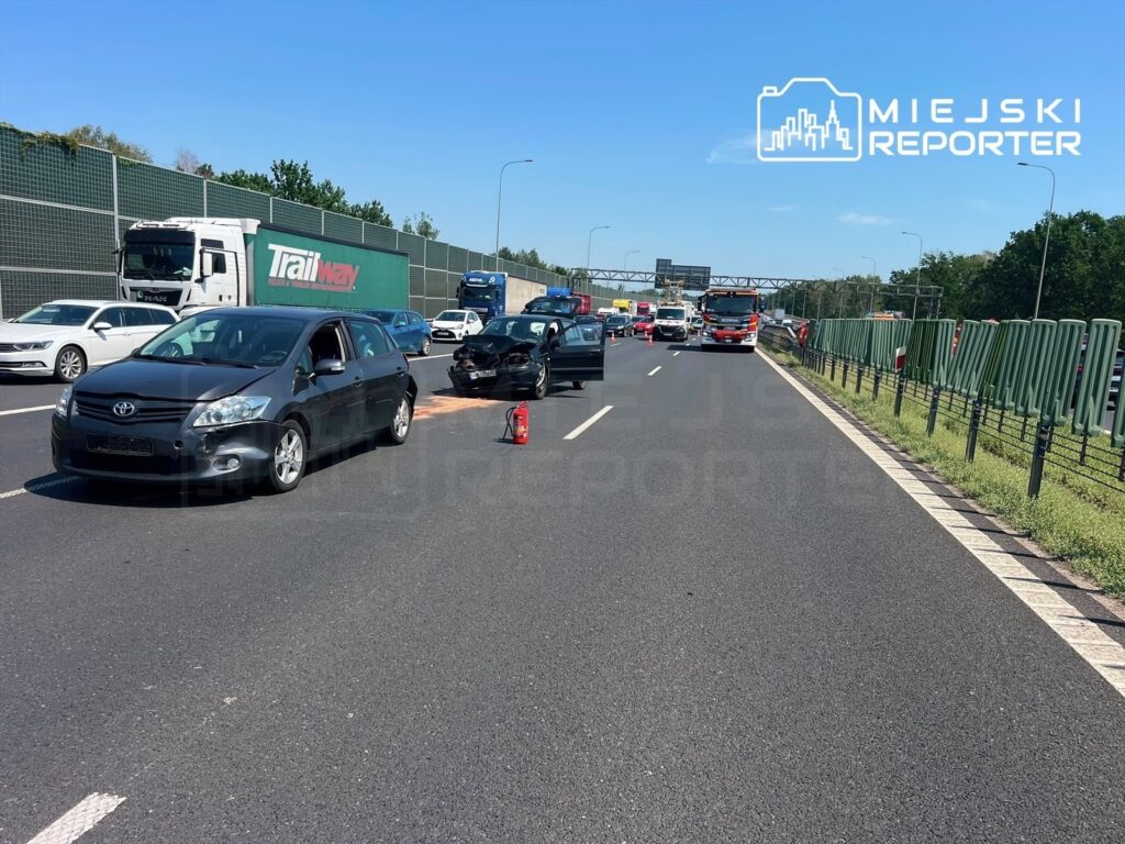 Czarny samochód osobowy zatrzymany na pasie ruchu, obok widoczny czerwony wóz strażacki, w tle ruchliwa autostrada.