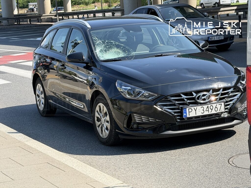 Czarny samochód osobowy z rozbitą szybą stoi na skrzyżowaniu ulic, obok pasów dla pieszych.
