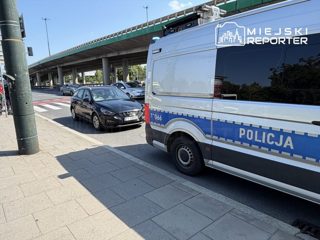 Policyjny radiowóz stojący na jezdni obok czarnego samochodu osobowego, w tle widoczna droga i zadaszenie.