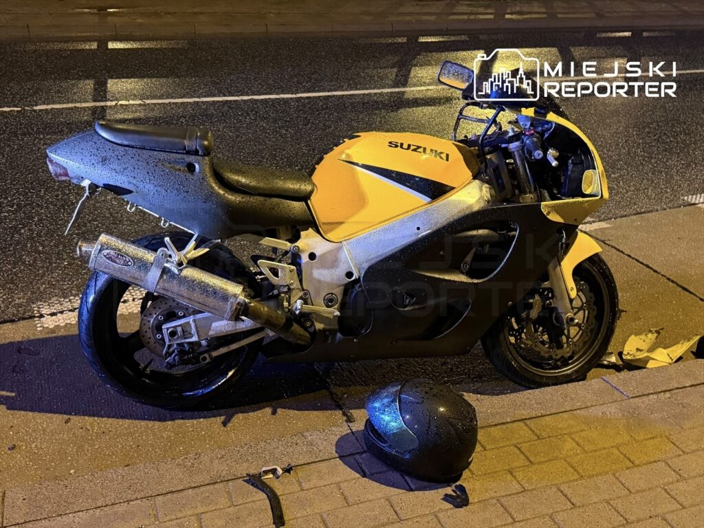 Motocykl marki Suzuki w żółto-szarej kolorystyce leży na poboczu drogi, obok znajduje się kask.