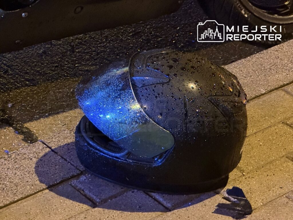 Czarny kask motocyklowy z kroplami wody leży na mokrej nawierzchni, obok zaparkowanego motocykla.
