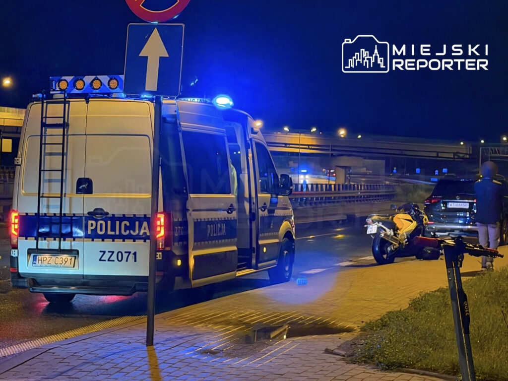 Policyjny radiowóz z włączonymi sygnałami świetlnymi zatrzymany na drodze obok przewróconego motocykla i samochodu osobowego.