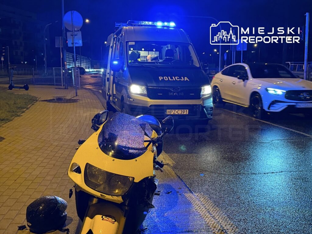 Żółty motocykl leży na drodze obok policyjnego pojazdu z włączonymi sygnalizatorami świetlnymi, a w tle widać inne samochody.