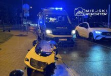 Żółty motocykl leży na drodze obok policyjnego pojazdu z włączonymi sygnalizatorami świetlnymi, a w tle widać inne samochody.