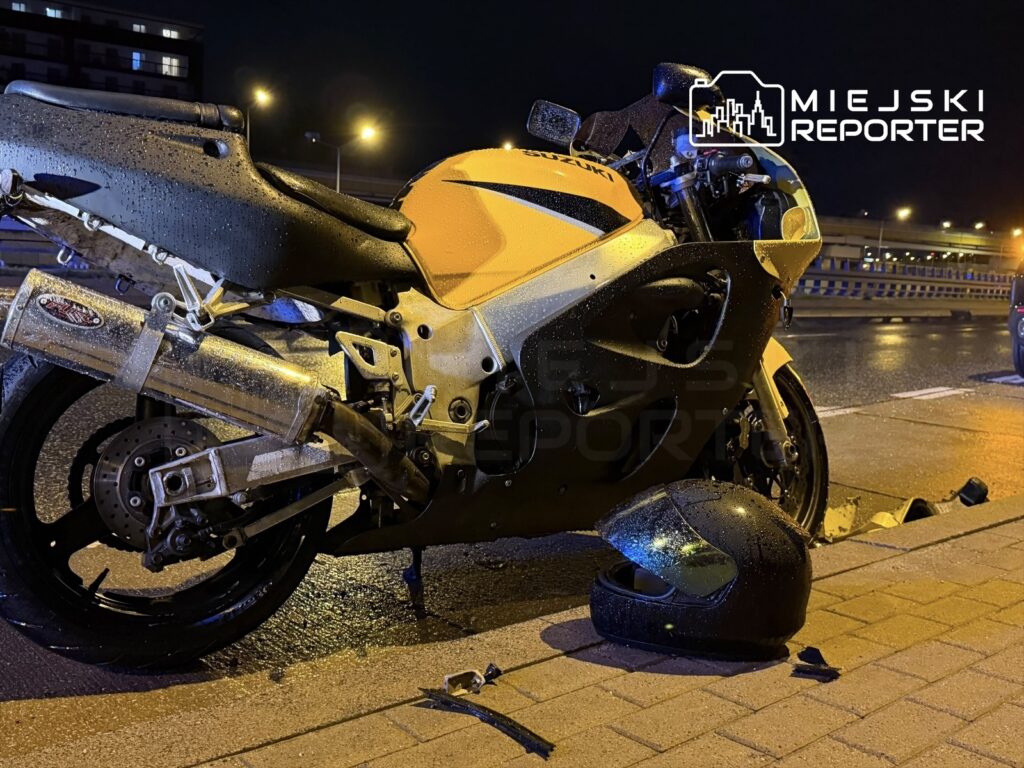 Zniszczony motocykl sportowy w deszczu, obok leży kask, na tle oświetlonej drogi nocą.