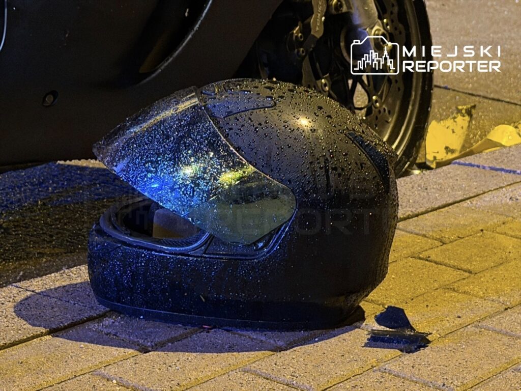 Czarny kask motocyklowy z kroplami wody leży na chodniku obok motocykla, w tle widoczne są policyjne taśmy.