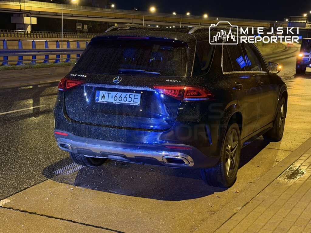 Czarny samochód marki Mercedes SUV zaparkowany na mokrej nawierzchni drogi nocą, z widocznymi kroplami deszczu.