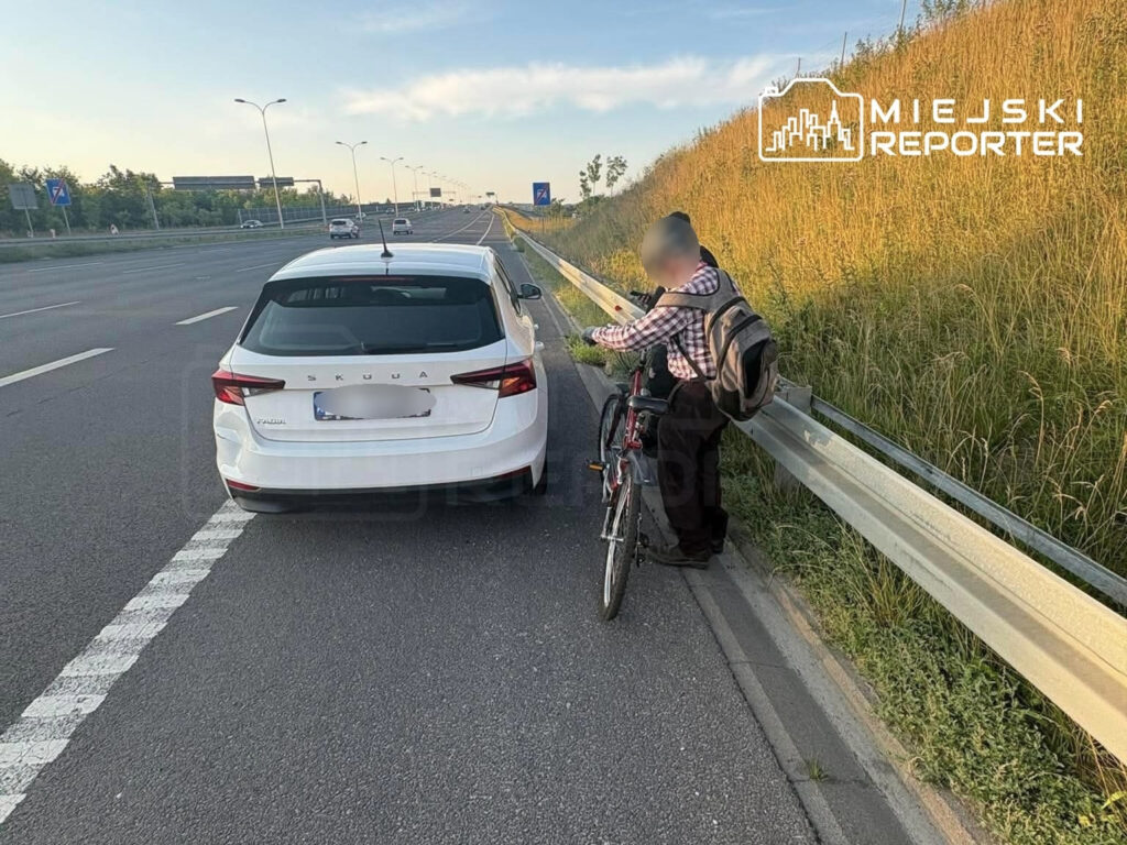 Starszy mężczyzna z plecakiem stoi obok roweru, rozmawiając z kierowcą zaparkowanej białej Skody na ekspresowej drodze.