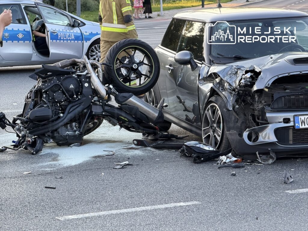 Motocykl leży na boku obok uszkodzonego samochodu, na drodze, z policjantami i strażakami w tle.