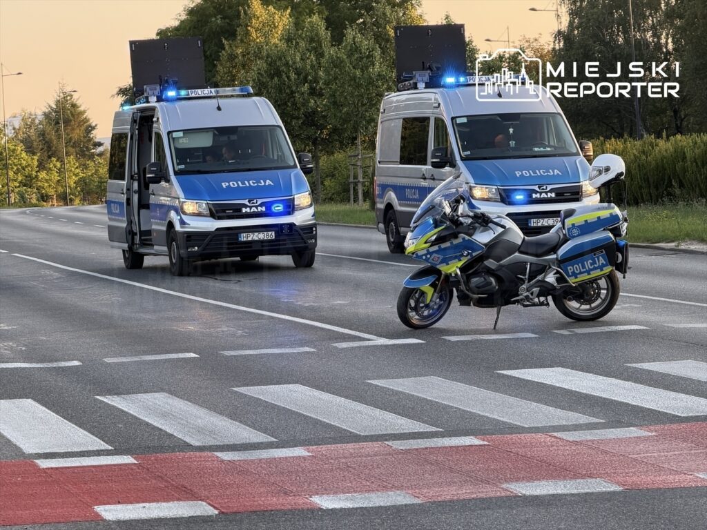 Policjanci w radiowozach i na motocyklu zabezpieczają miejsce zdarzenia na drodze.