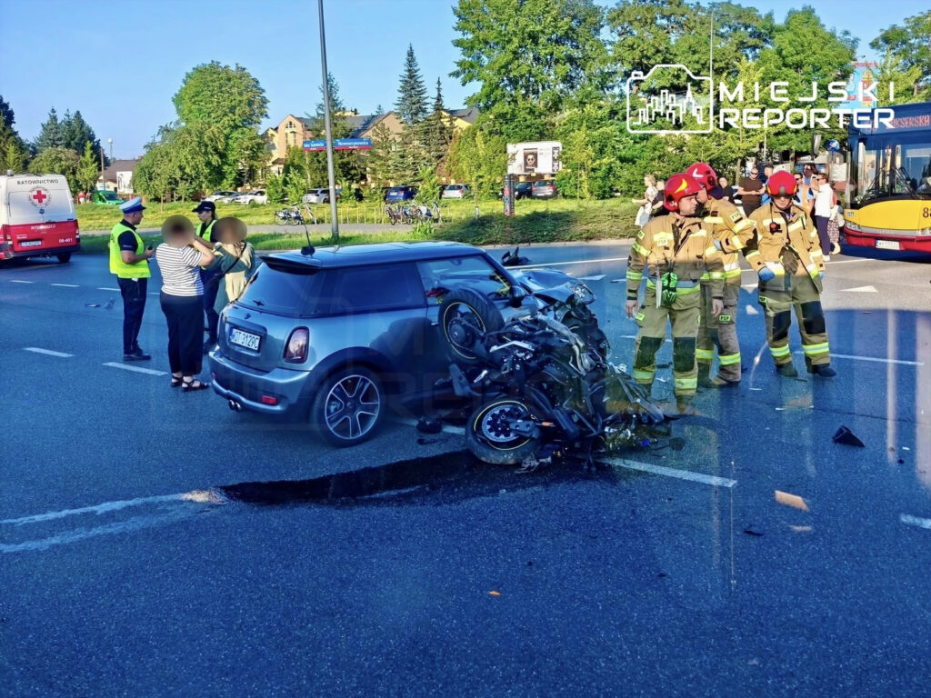 Motocyklista leży obok uszkodzonego motocykla, a strażacy oraz policjant rozmawiają z osobami przy Mini.