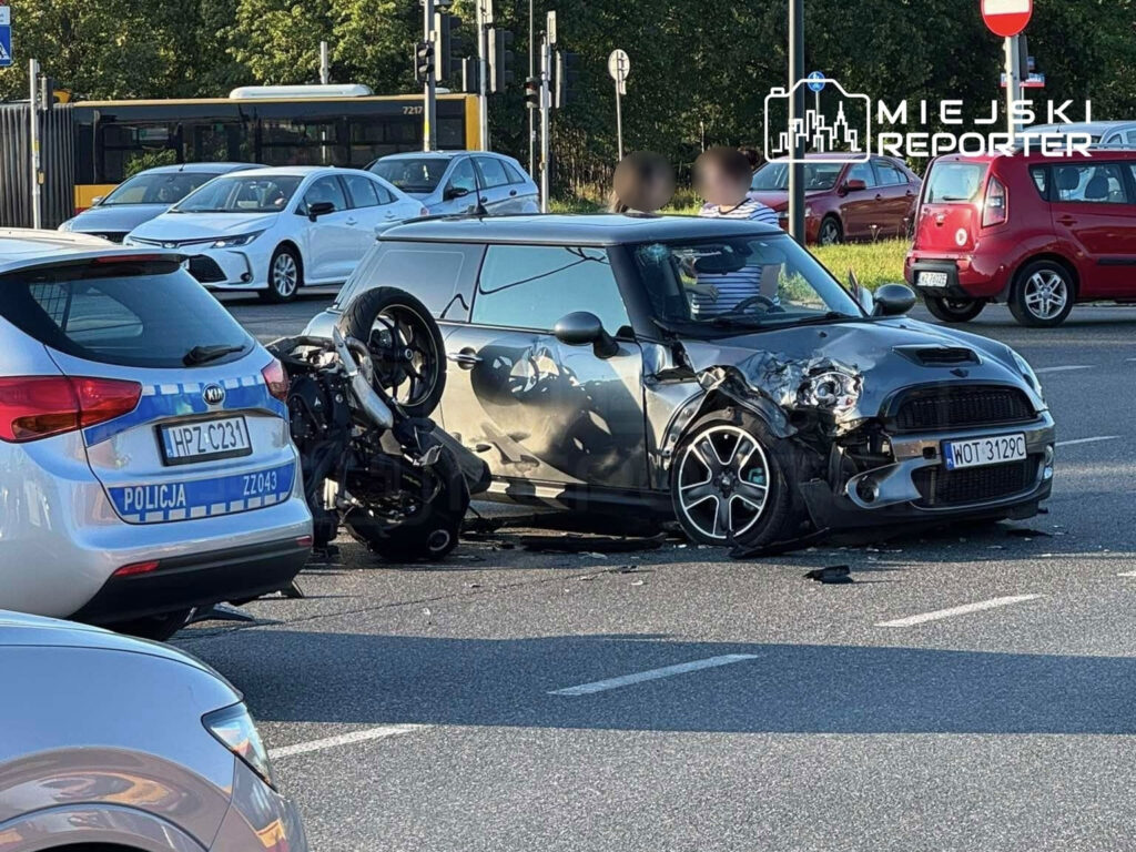 Motocykl leży na boku obok uszkodzonego samochodu Mini na ruchliwej ulicy, a w tle widać inne pojazdy.