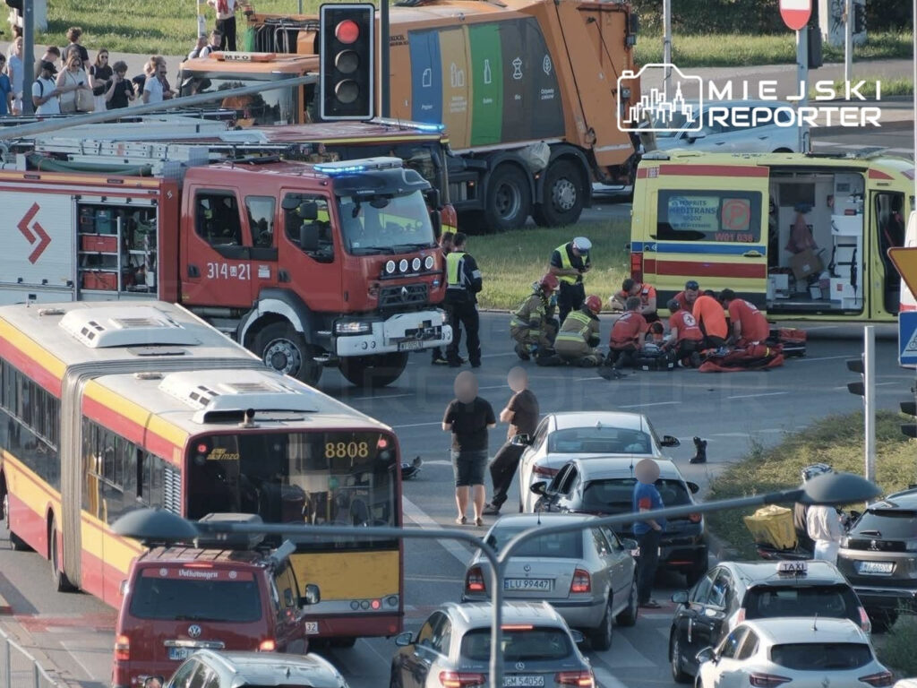 Zespół ratunkowy udziela pomocy poszkodowanemu motocykliście na skrzyżowaniu, obok czerwonego wozu strażackiego.