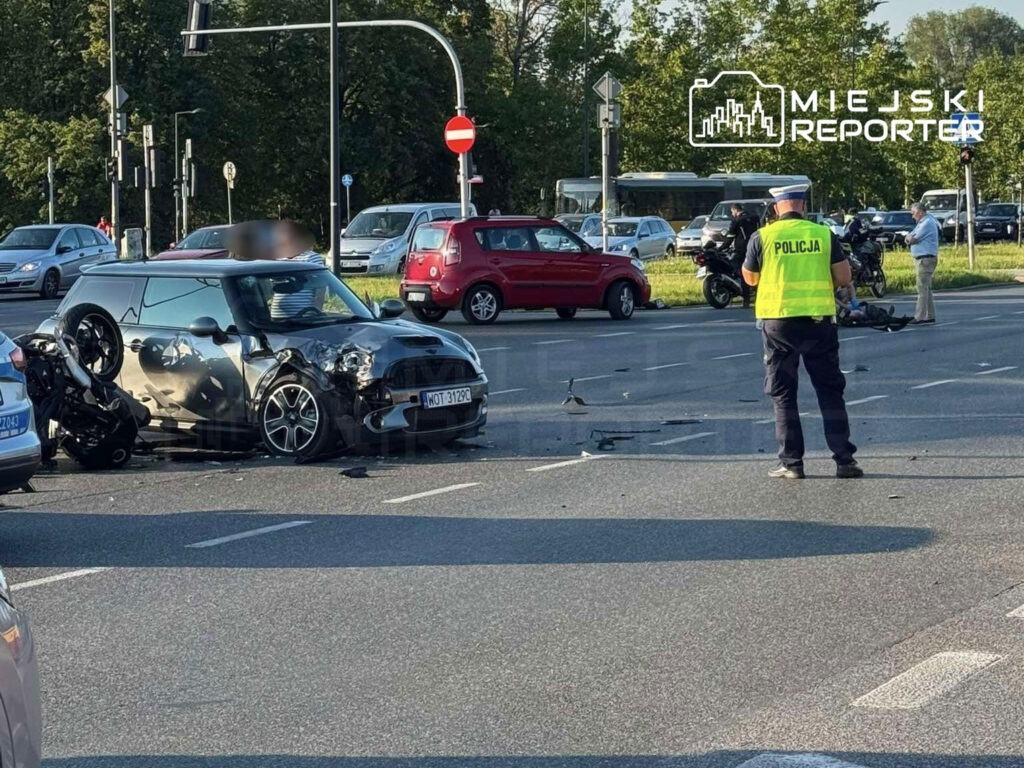 W zderzeniu motocyklowym z samochodem Mini, uszkodzona maszyna leży na boku, a policjant zabezpiecza miejsce wypadku.