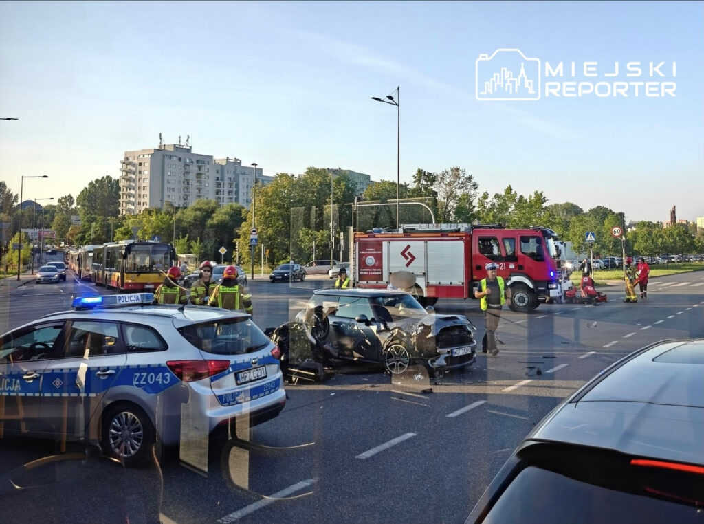 Na skrzyżowaniu stoją policjanci i strażacy, obok zderzone auta, w tym uszkodzony motocykl. W tle widać autobusy i budynki.