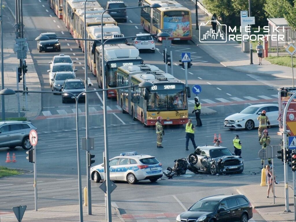 Na skrzyżowaniu stoi rozbity motocykl obok uszkodzonego samochodu osobowego, a w tle widać ruchliwe ulice z autobusami.