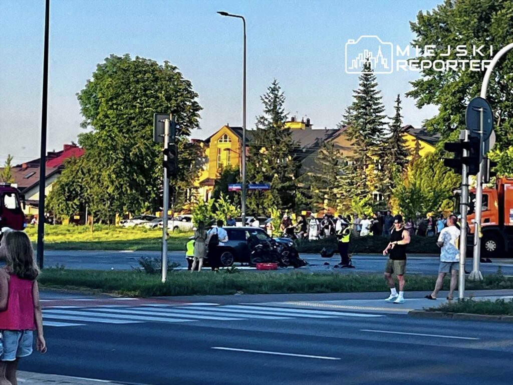 Motocyklista leży na jezdni obok uszkodzonego motocykla, a w tle gromadzą się świadkowie zdarzenia.