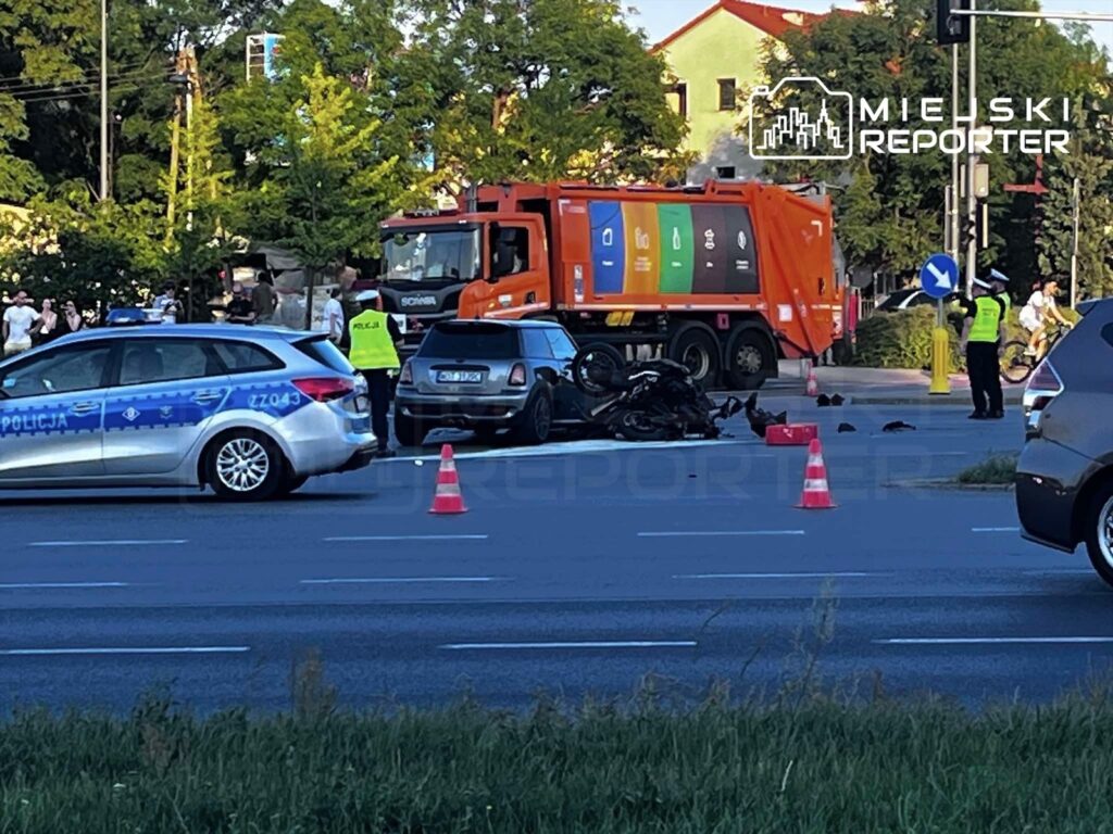 Motocyklista leży na jezdni po wypadku z samochodem, obok stoją policjanci i zaparkowane pojazdy.