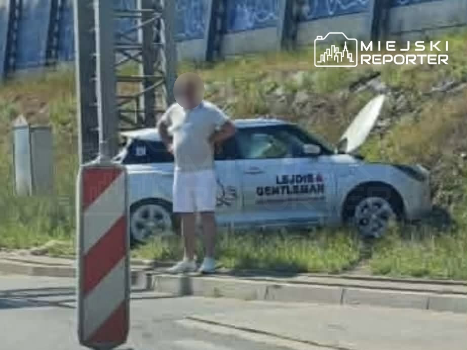 Mężczyzna w szortach i koszulce stoi obok samochodu nauki jazdy z otwartą maską, przy drodze.