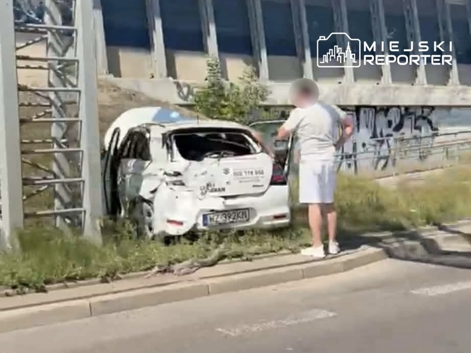Zniszczony samochód nauki jazdy stoi na poboczu drogi, a mężczyzna w jasnej odzieży sprawdza jego stan.