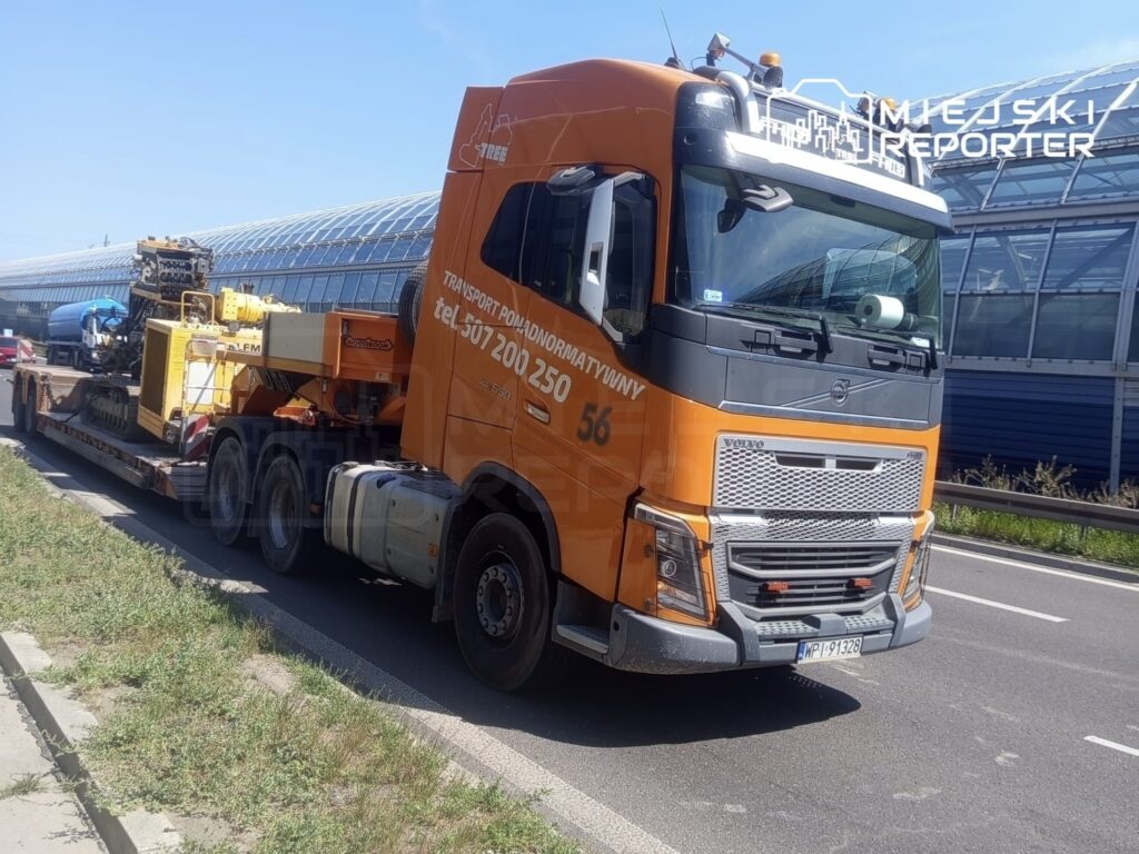 Czterokołowa ciężarówka z pomarańczową kabiną transportującą sprzęt budowlany na przyczepie, zatrzymana na drodze.