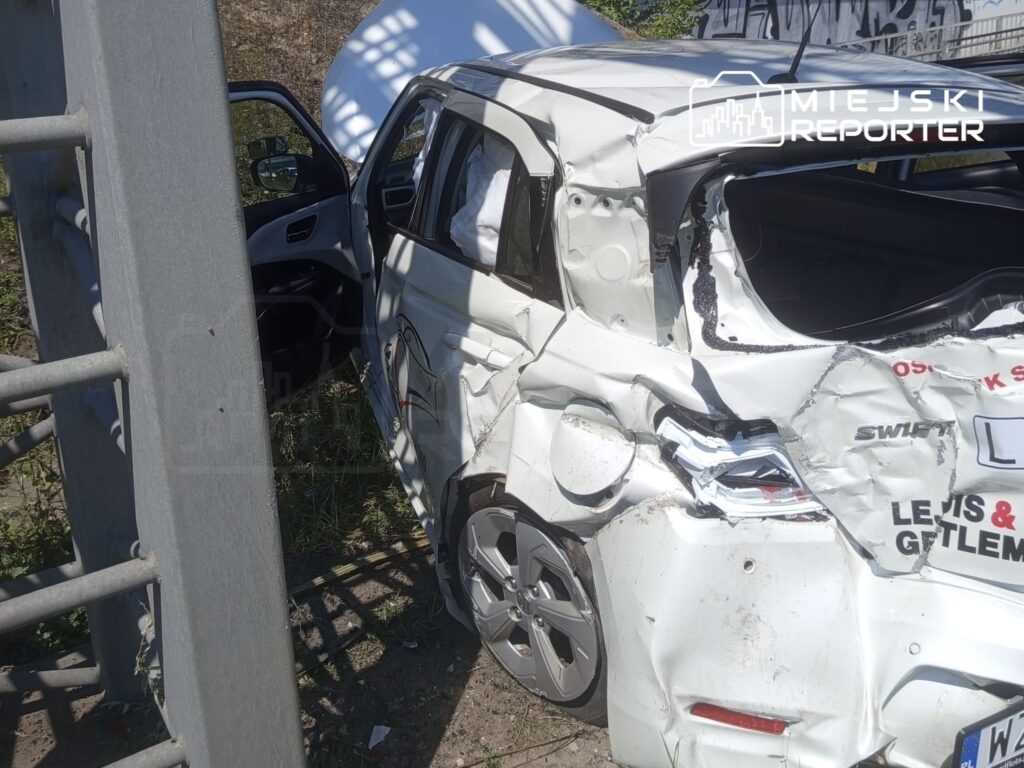 Zniszczony samochód nauki jazdy z wygiętą karoserią stoi obok metalowego słupa, otoczony trawą i graffiti.