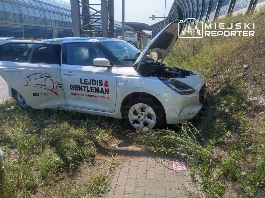 Uszkodzony samochód nauki jazdy z otwartą maską stoi na poboczu drogi, otoczony wysoką trawą.