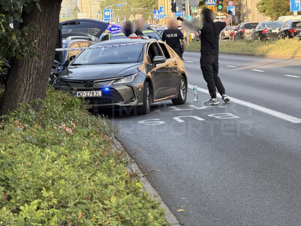Mężczyzna w czarnych ubraniach stoi z uniesionymi rękami obok zatrzymanej Toyoty, przy drodze w mieście. Policjanci zabezpieczają miejsce zdarzenia.
