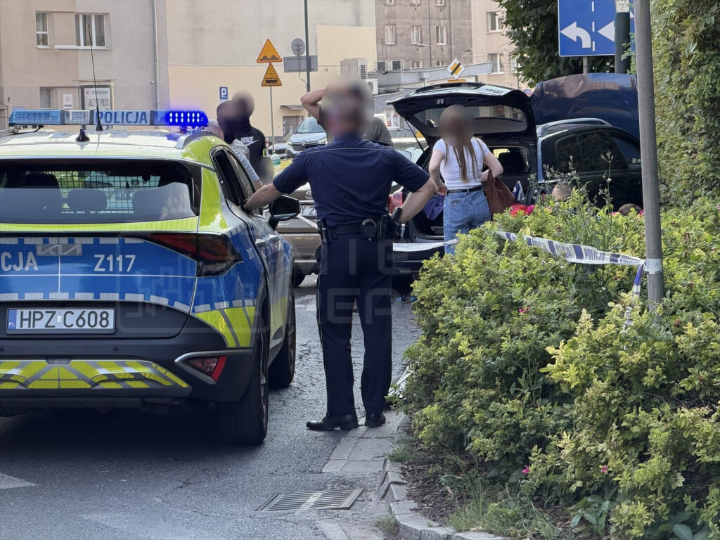 Policjant rozmawia z mężczyzną obok radiowozu, podczas gdy inne osoby przeszukują bagażnik samochodu.