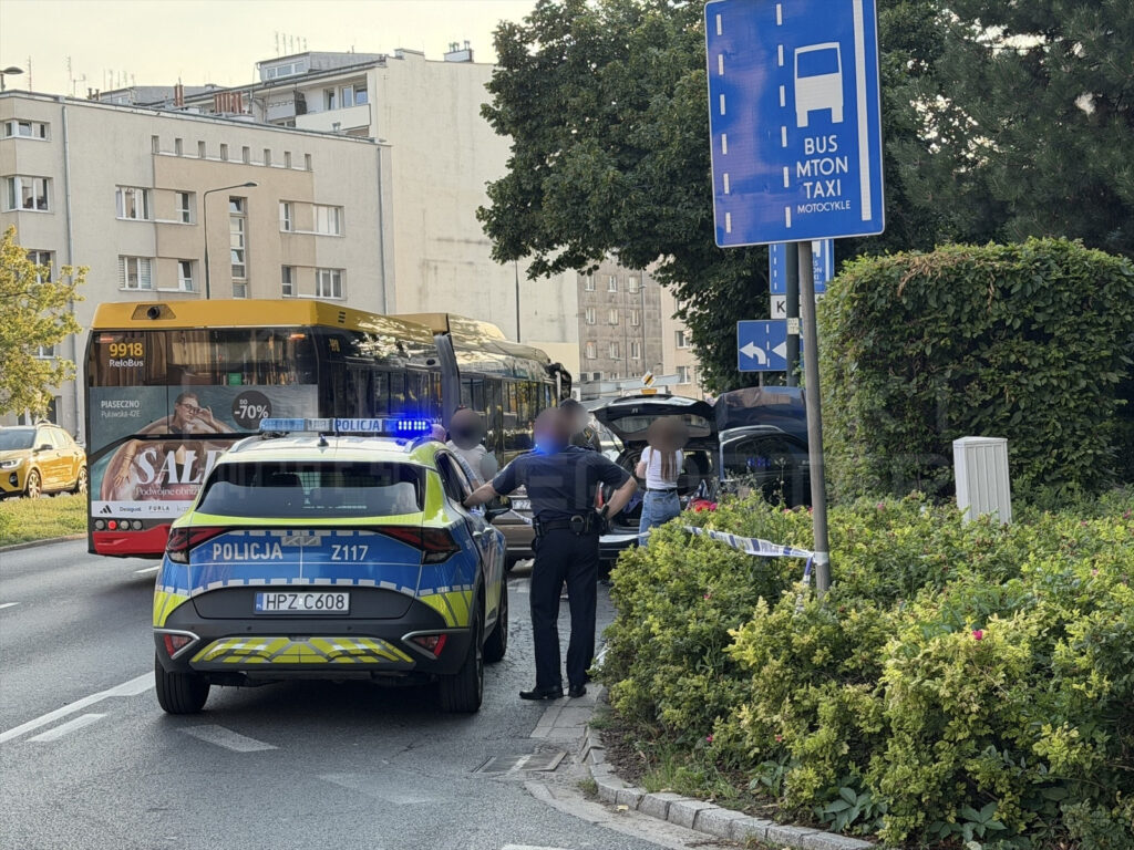 Policyjny radiowóz z włączonymi sygnałami stoi przy drodze, obok otwartego bagażnika samochodu. W tle autobus komunikacji miejskiej.