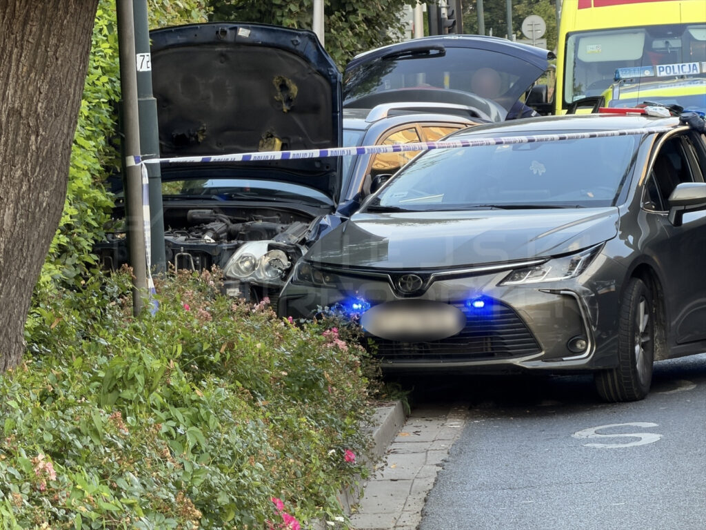 Czarny samochód z otwartą maską stoi obok latarni, a obok niego policyjny pojazd z włączonymi sygnałami świetlnymi.
