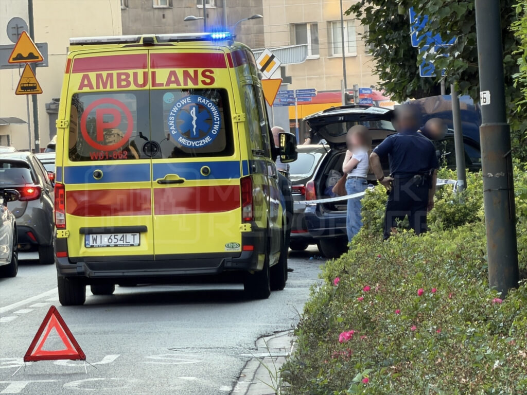 Ambulans z włączonymi sygnałami świetlnymi zatrzymany na ulicy, obok stoi policjant rozmawiający z osobą przy otwartym bagażniku.