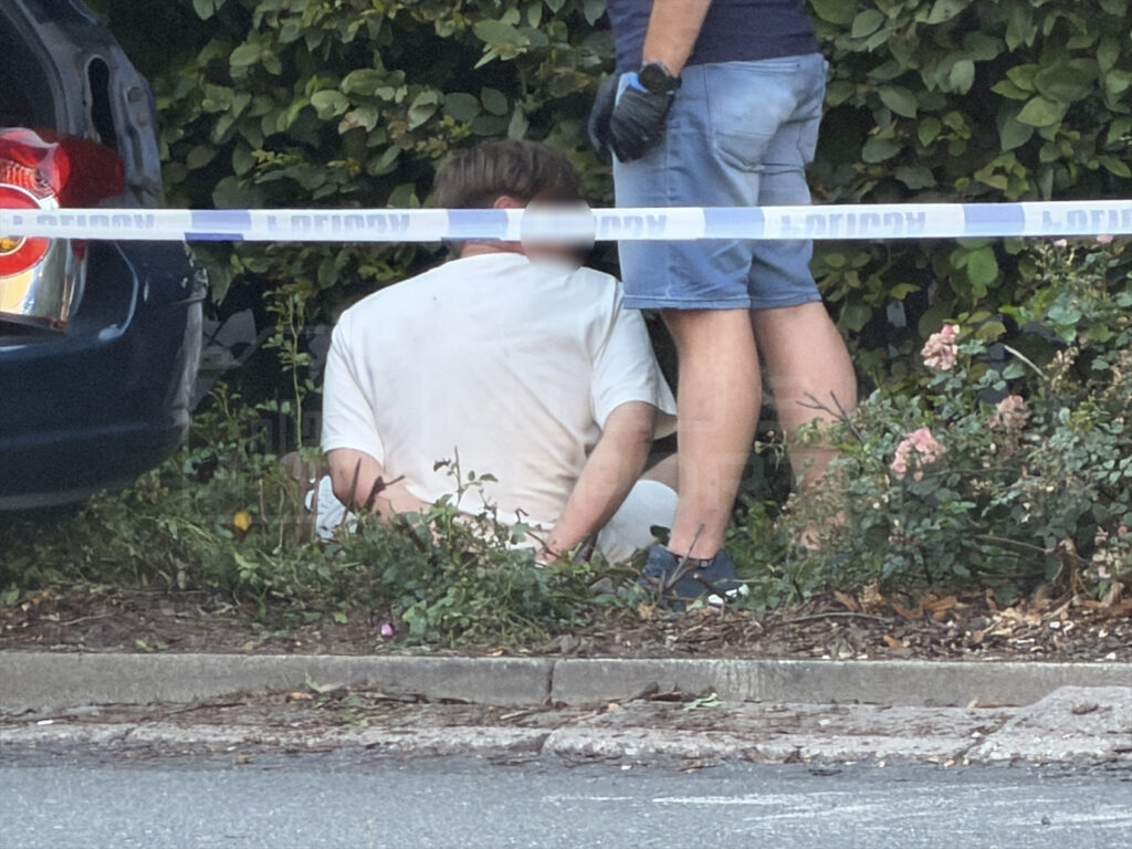 Mężczyzna w białej koszuli siedzący na ziemi, a obok niego funkcjonariusz w niebieskiej koszulce, w otoczeniu zieleni.