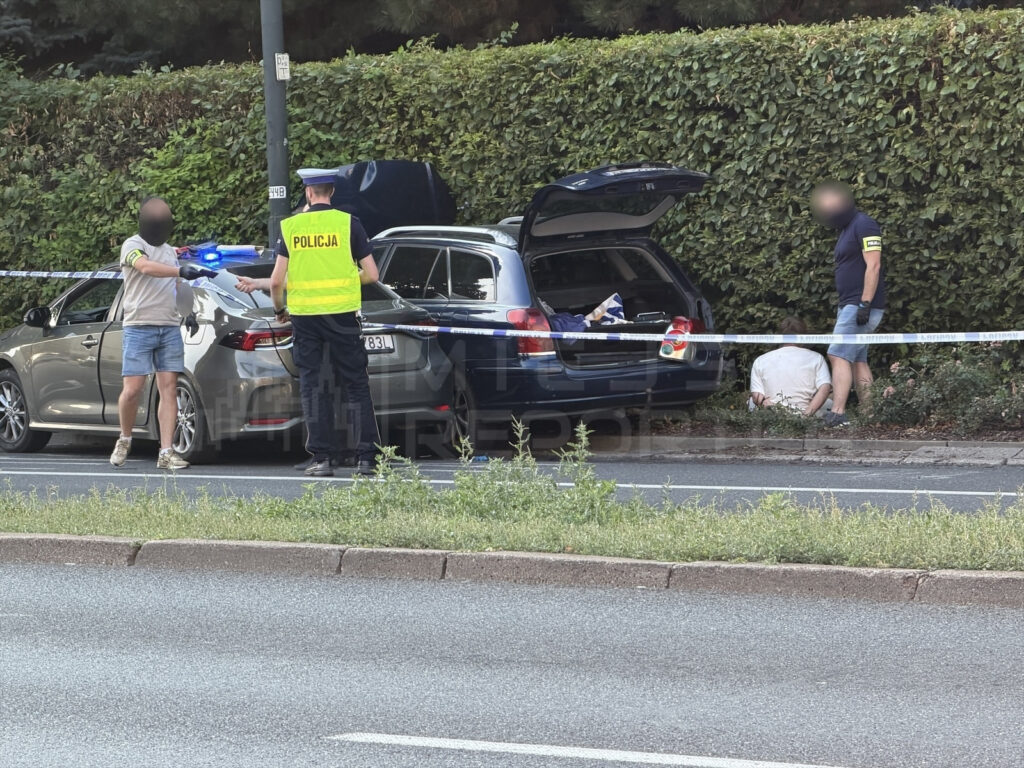 Policjant w kamizelce odblaskowej rozmawia z mężczyzną w krótkich spodniach obok zatrzymanego samochodu.
