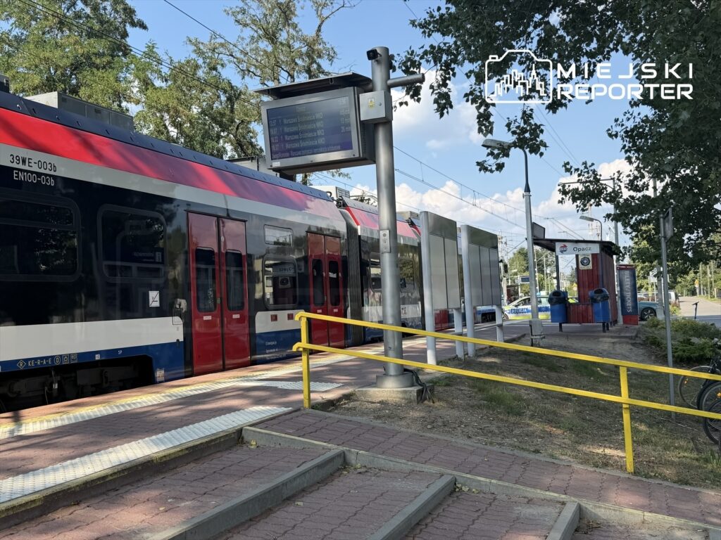 Pociąg osobowy zatrzymany na stacji, obok widoczny rozkład jazdy oraz przystanek autobusowy w otoczeniu zieleni.