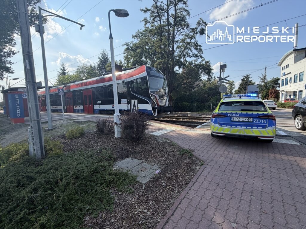 Tramwaj zatrzymany na przystanku obok radiowozu policyjnego z włączonymi sygnałami świetlnymi.