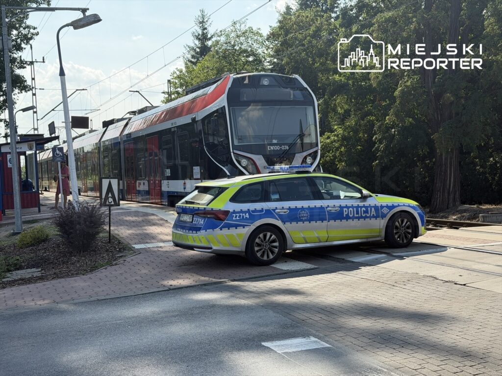 Policjant w radiowozie zatrzymuje ruch na torowisku tramwajowym obok przystanku w słoneczny dzień.