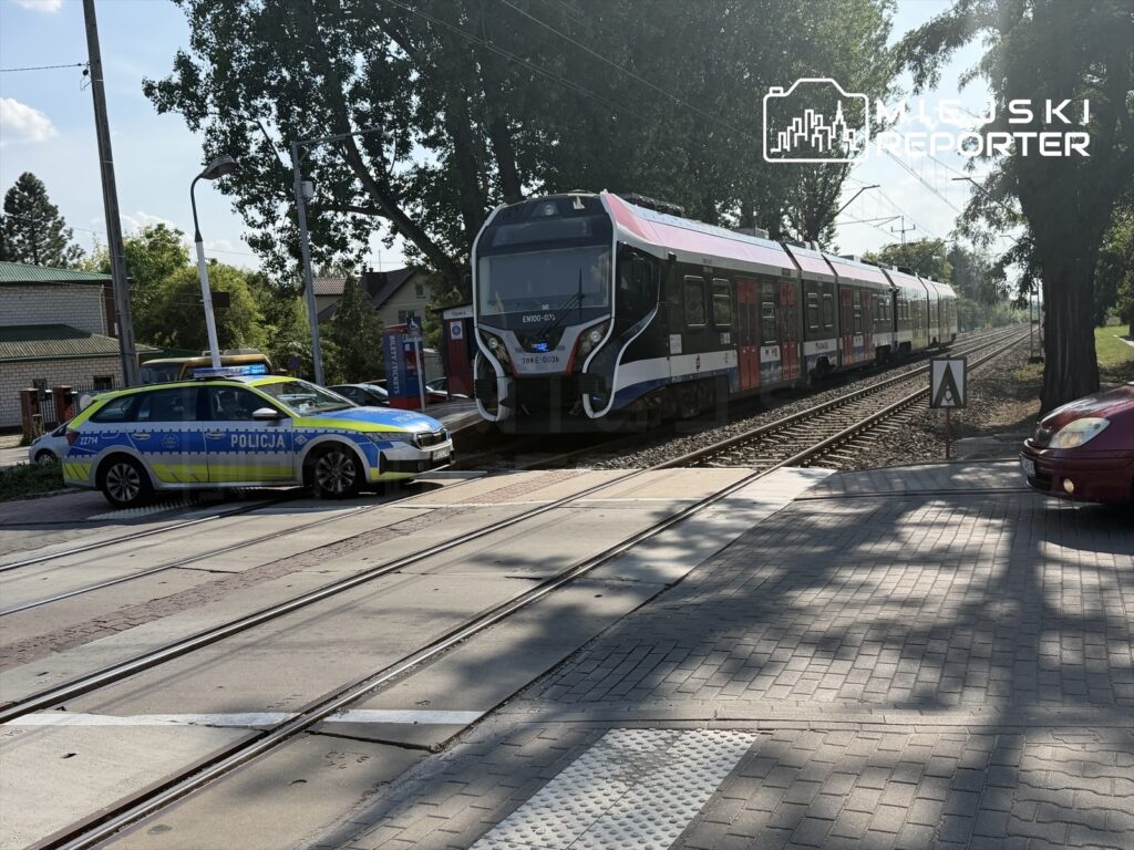 Policjant w radiowozie stoi obok torów kolejowych, obok przejeżdżającego pociągu regionalnego.