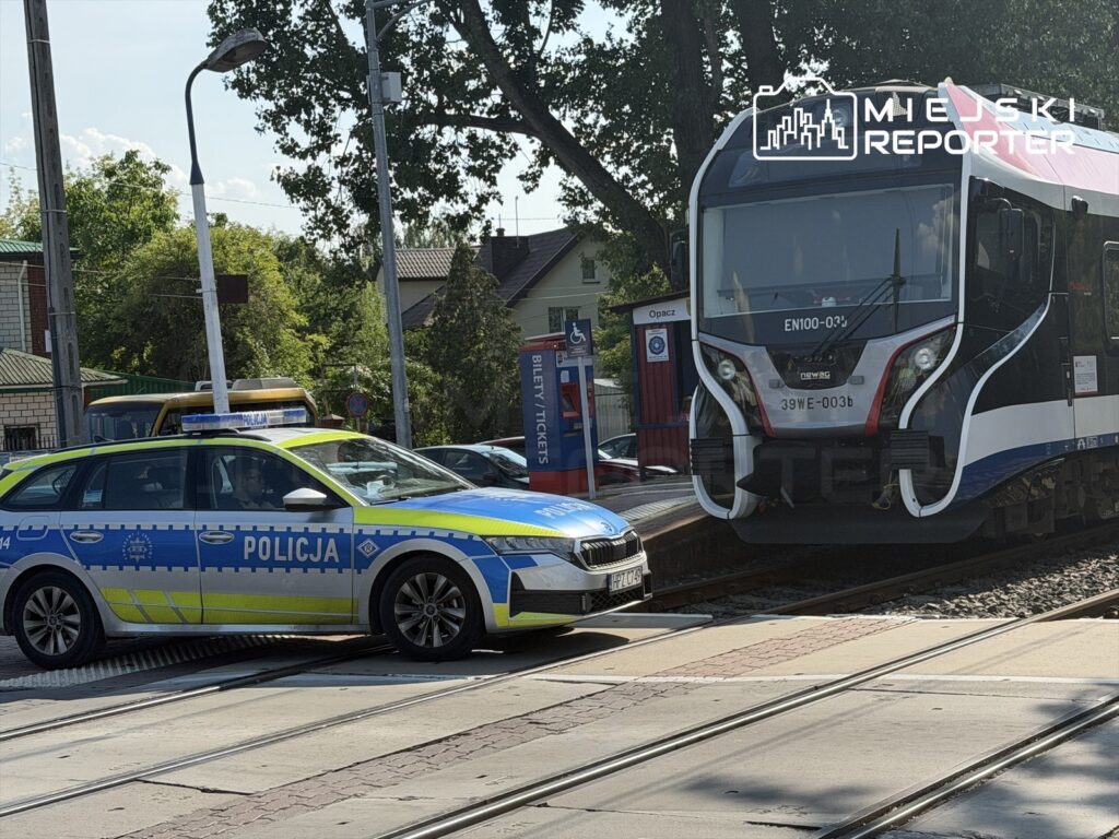Policyjny radiowóz zatrzymany obok torów, obok nowoczesnego pociągu na stacji kolejowej w słoneczny dzień.