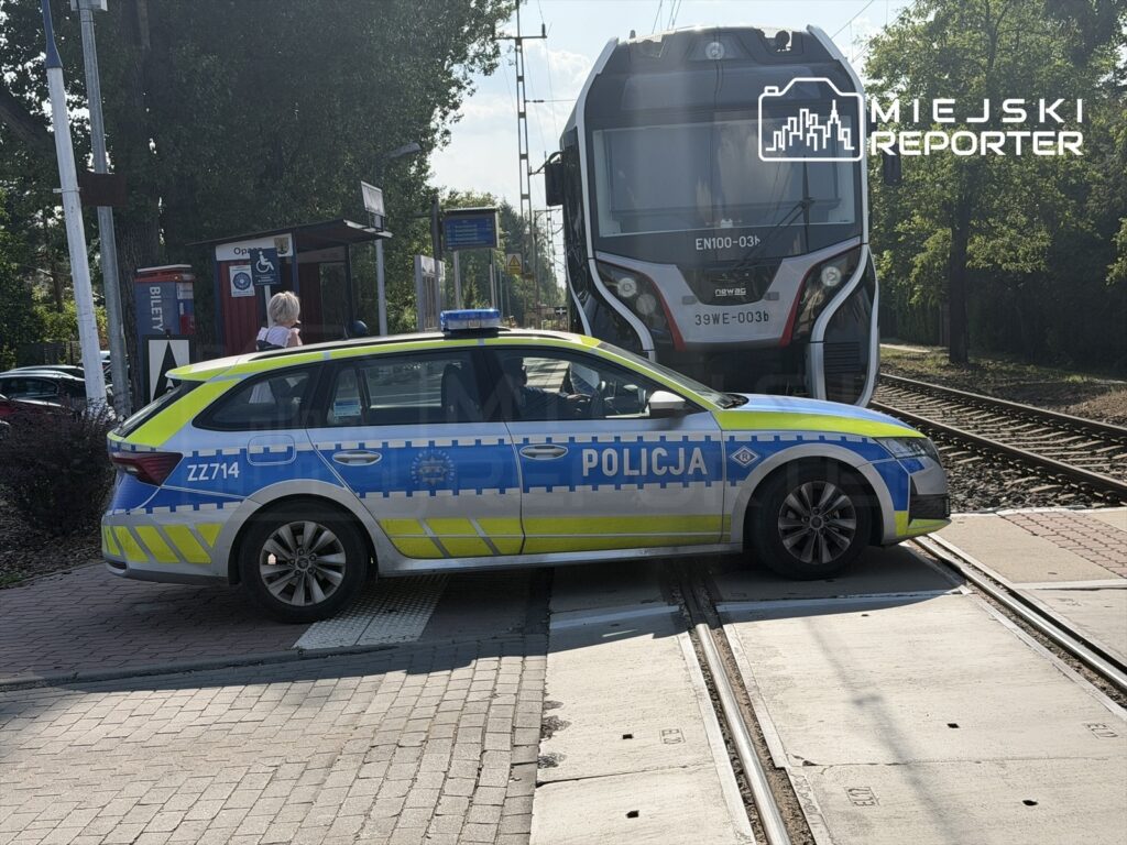 Policyjny radiowóz stoi na torach kolejowych obok nadjeżdżającego pociągu, w tle widoczny przystanek.