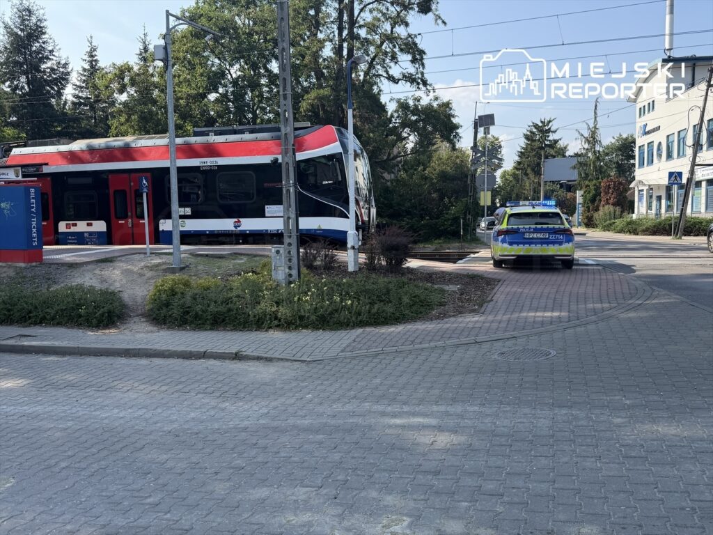 Tramwaj zatrzymany na przystanku obok stojącego radiowozu policyjnego w miejskim otoczeniu.