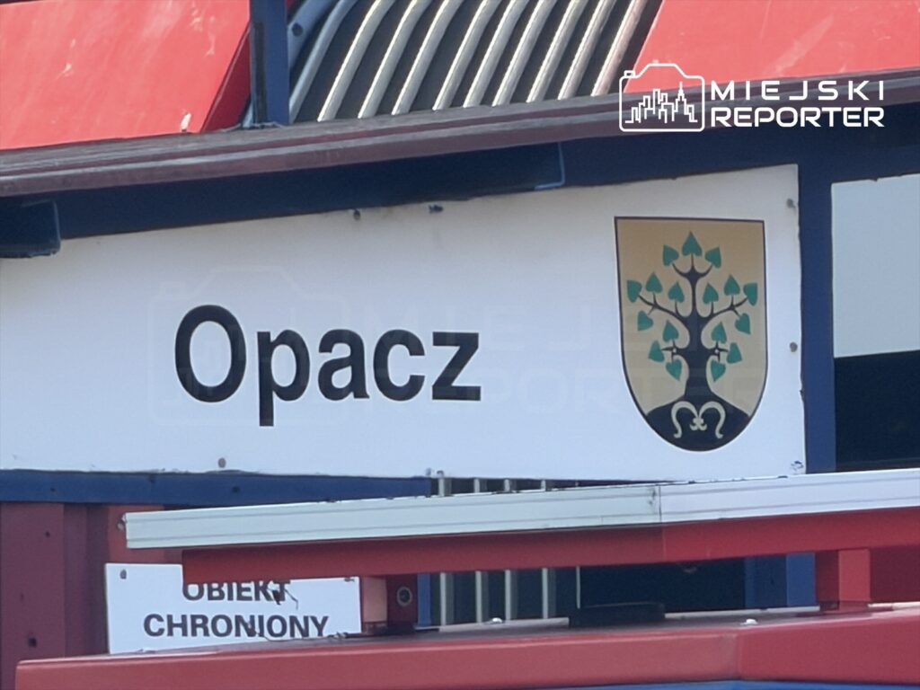 Tablica informacyjna z napisem "Opacz" oraz herbem przedstawiającym drzewo na tle złotym, umieszczona na budynku.